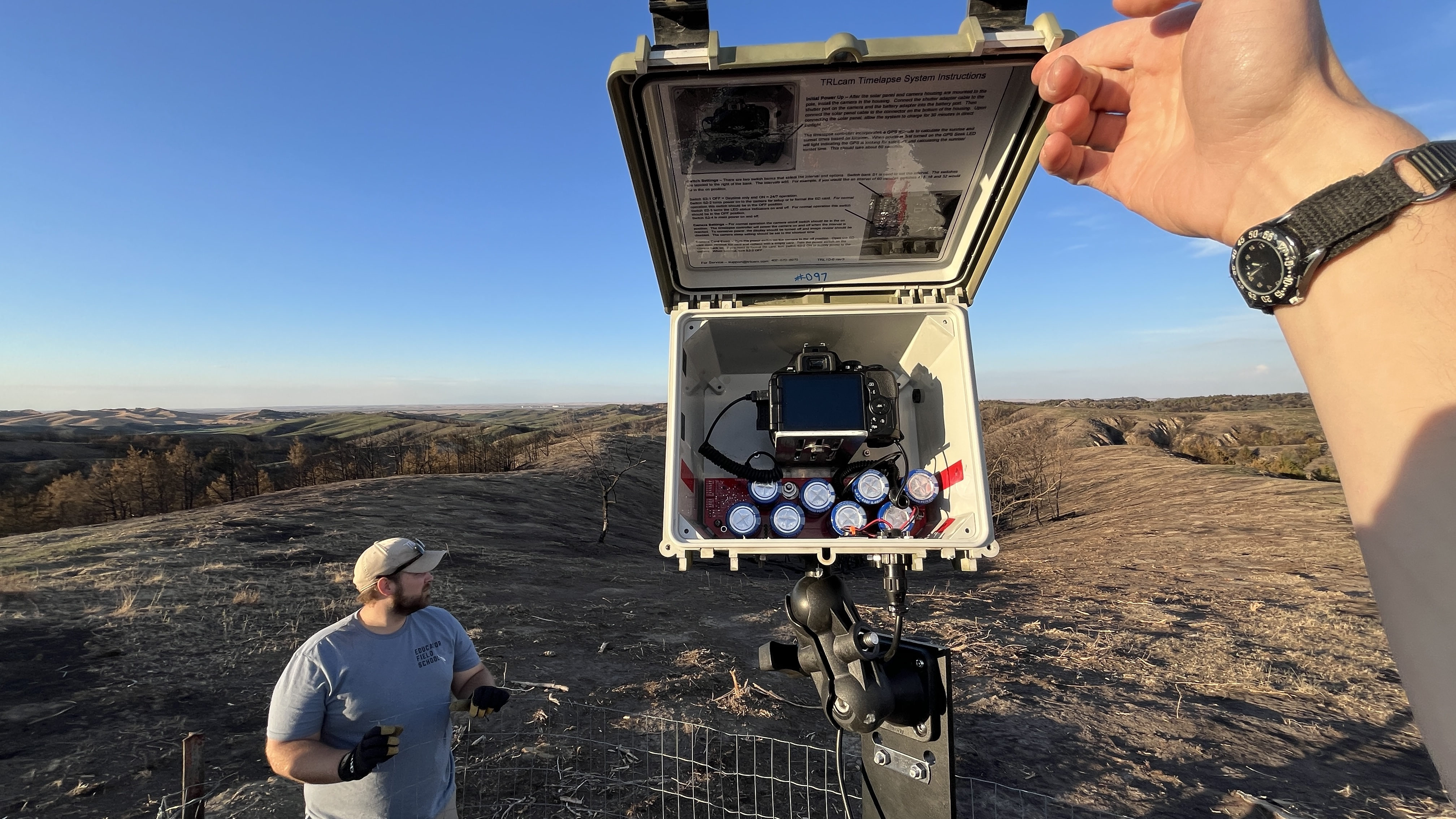 Open equipment box with camera and batteries; person standing in a grassy, hilly landscape at sunset.