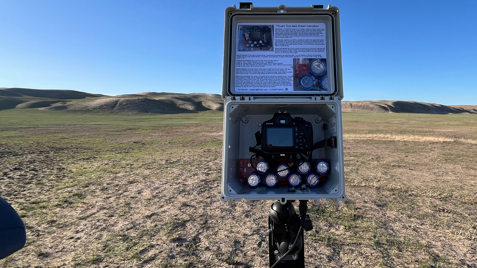 Open equipment box on a tripod in a grassy, hilly outdoor setting under a clear blue sky.